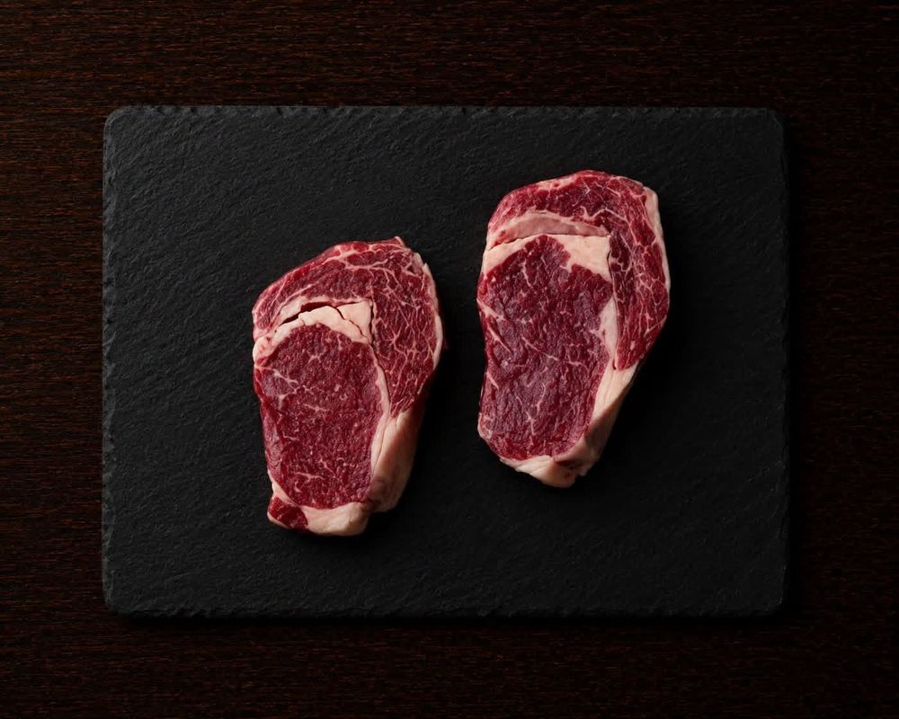 Two raw ribeye steaks on a dark slate board