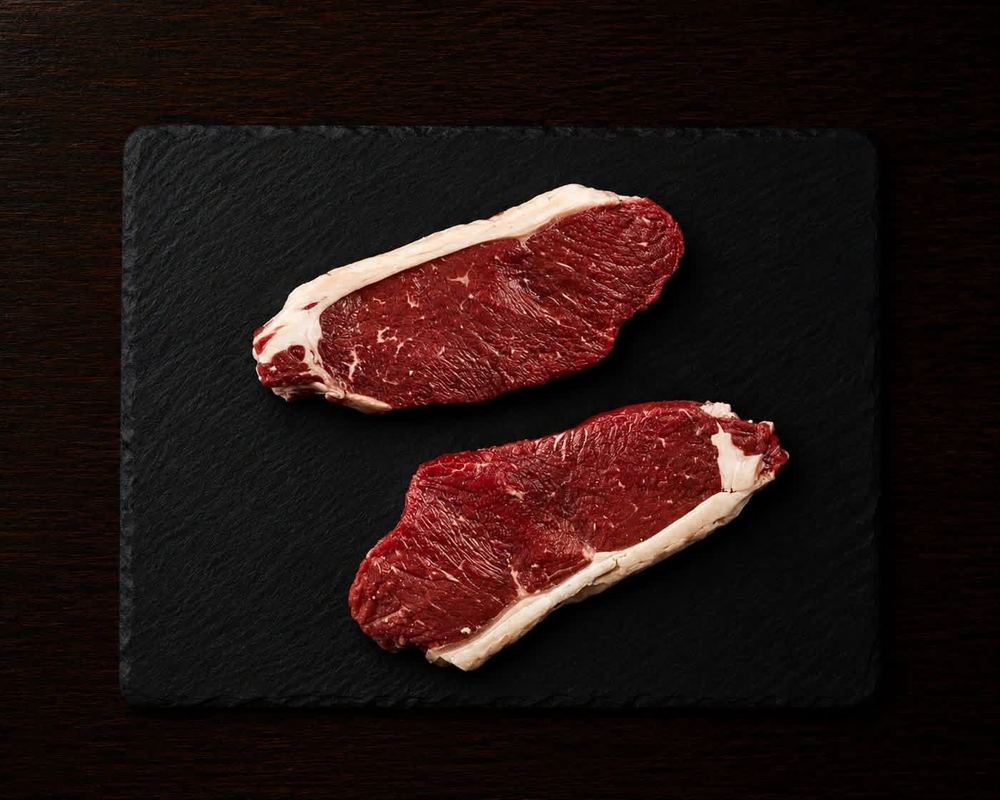 Two raw New York strip steaks on a dark slate board